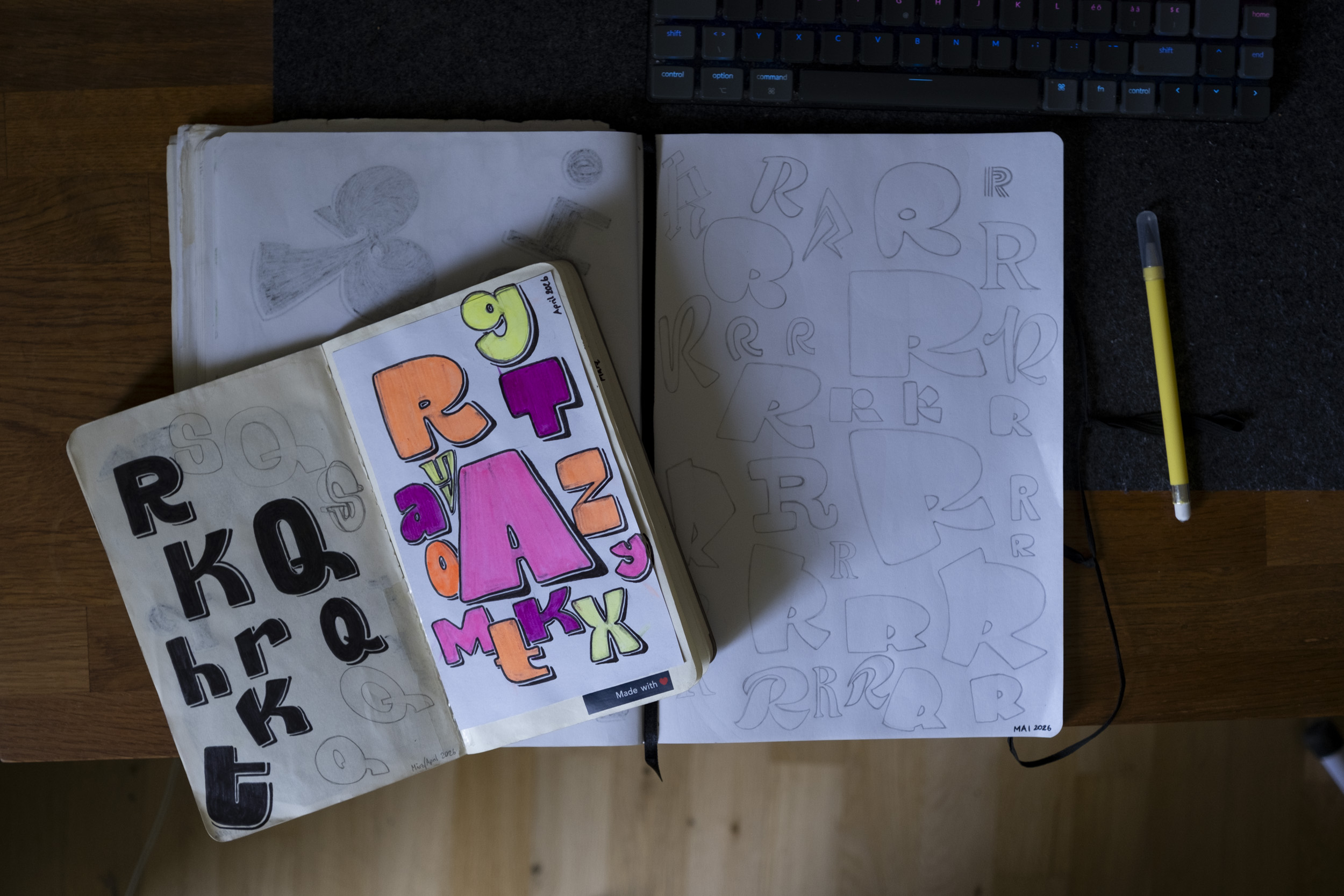 Sketchbooks open on a wooden desk showing hand-drawn lettering practice — bold black bubble letters on the left, colorful marker-filled letters on a middle insert, and pencil-sketched variations of the letter "R" on the right.