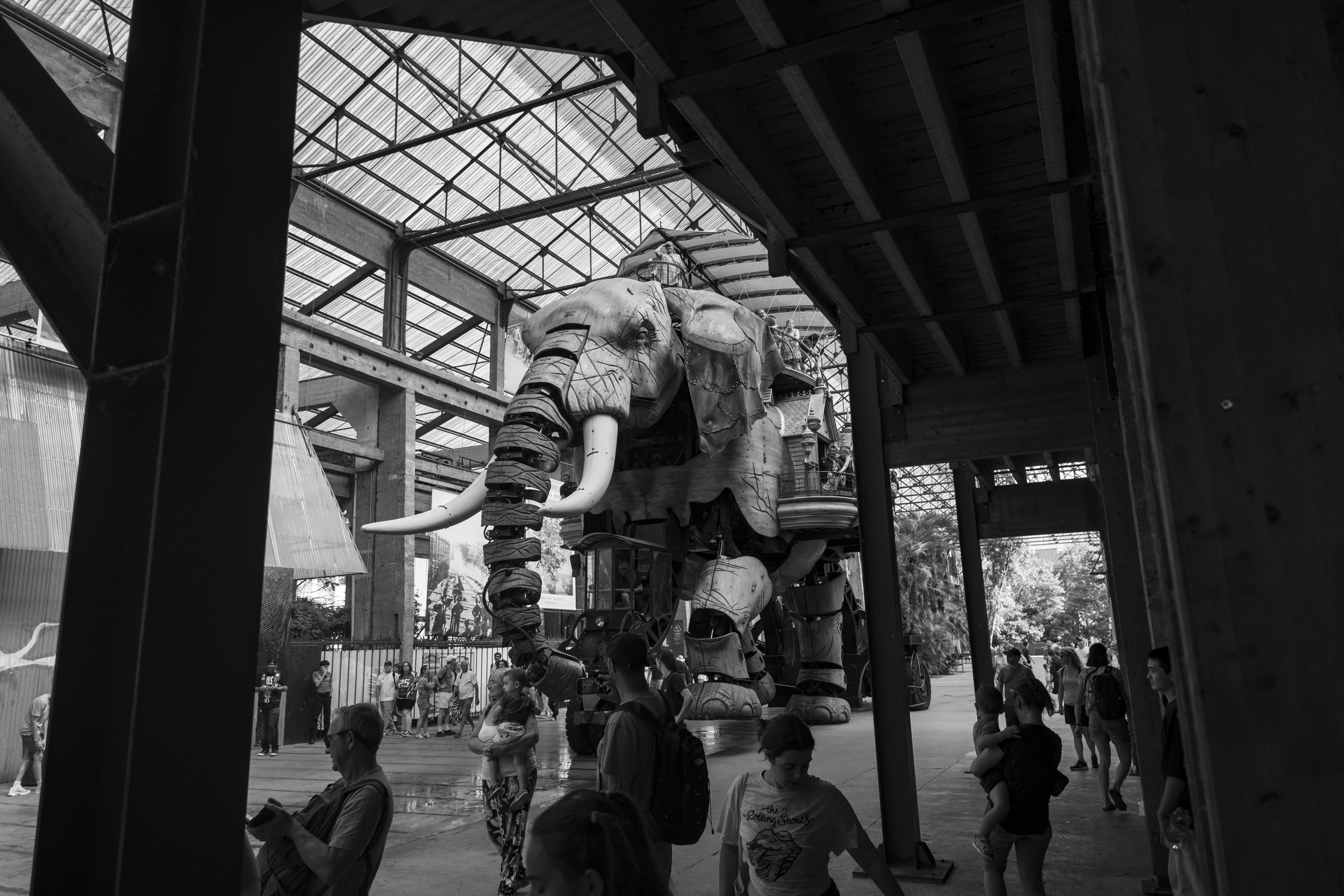 Black-and-white photo of the Grand Éléphant at Les Machines de l'île in Nantes — a giant mechanical elephant built of wood and steel, with long tusks and an ornate multi-level carriage on its back carrying visitors. It stands under the roof of the former shipyard hall, with a crowd of people walking and watching in the foreground.