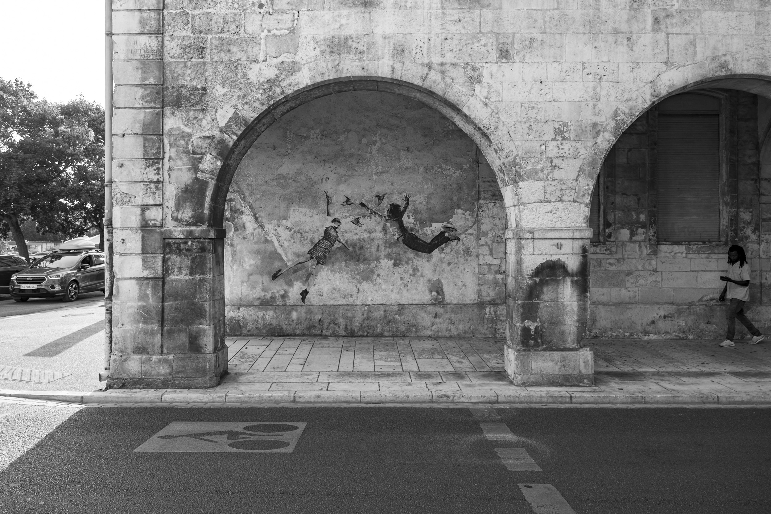 Black-and-white street photo of a mural showing two floating figures and birds under a stone arch, with a person walking past.