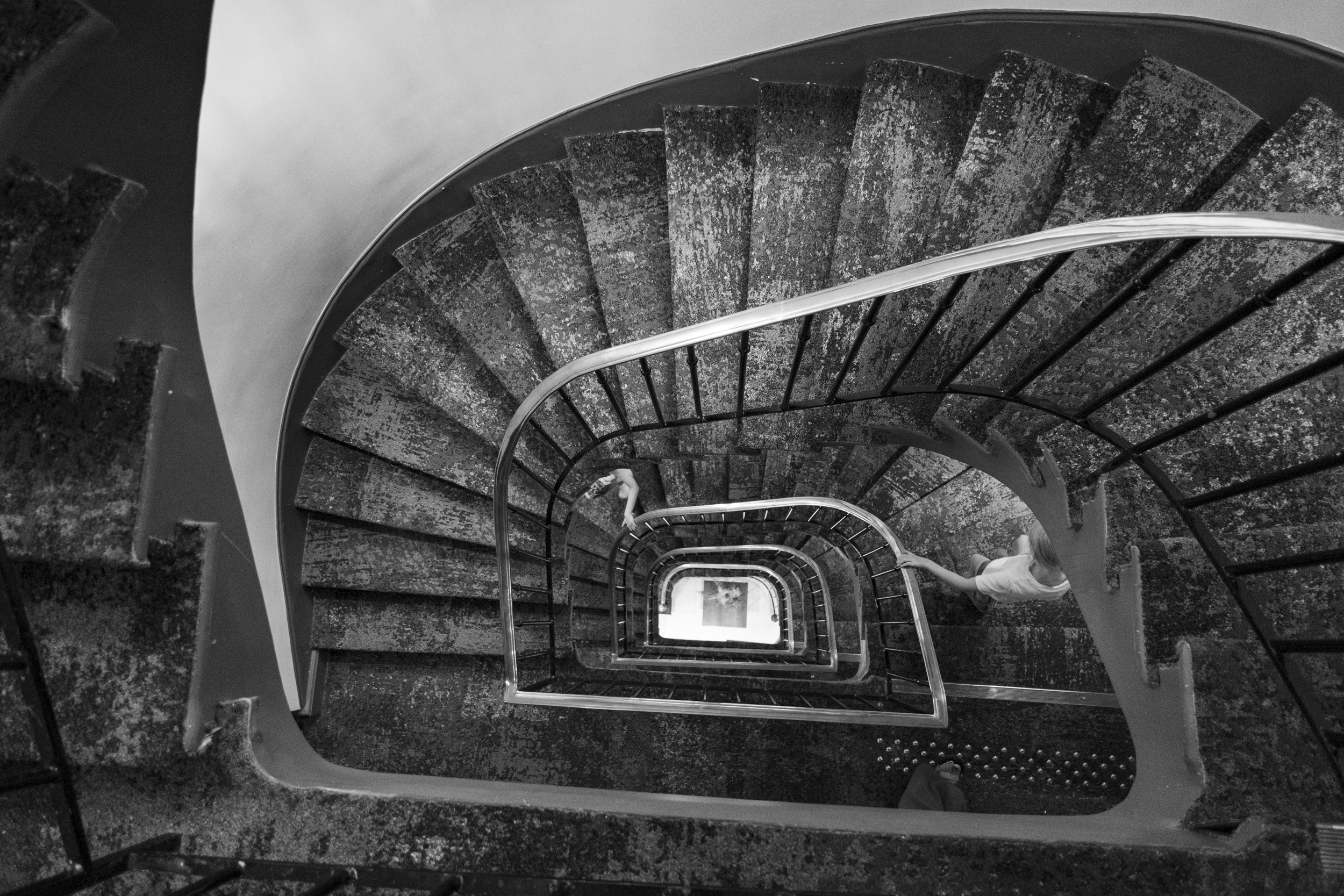 Black-and-white view down the multi-story spiral staircase of our hotel, with the kids walking down the stairs, holding the railings.