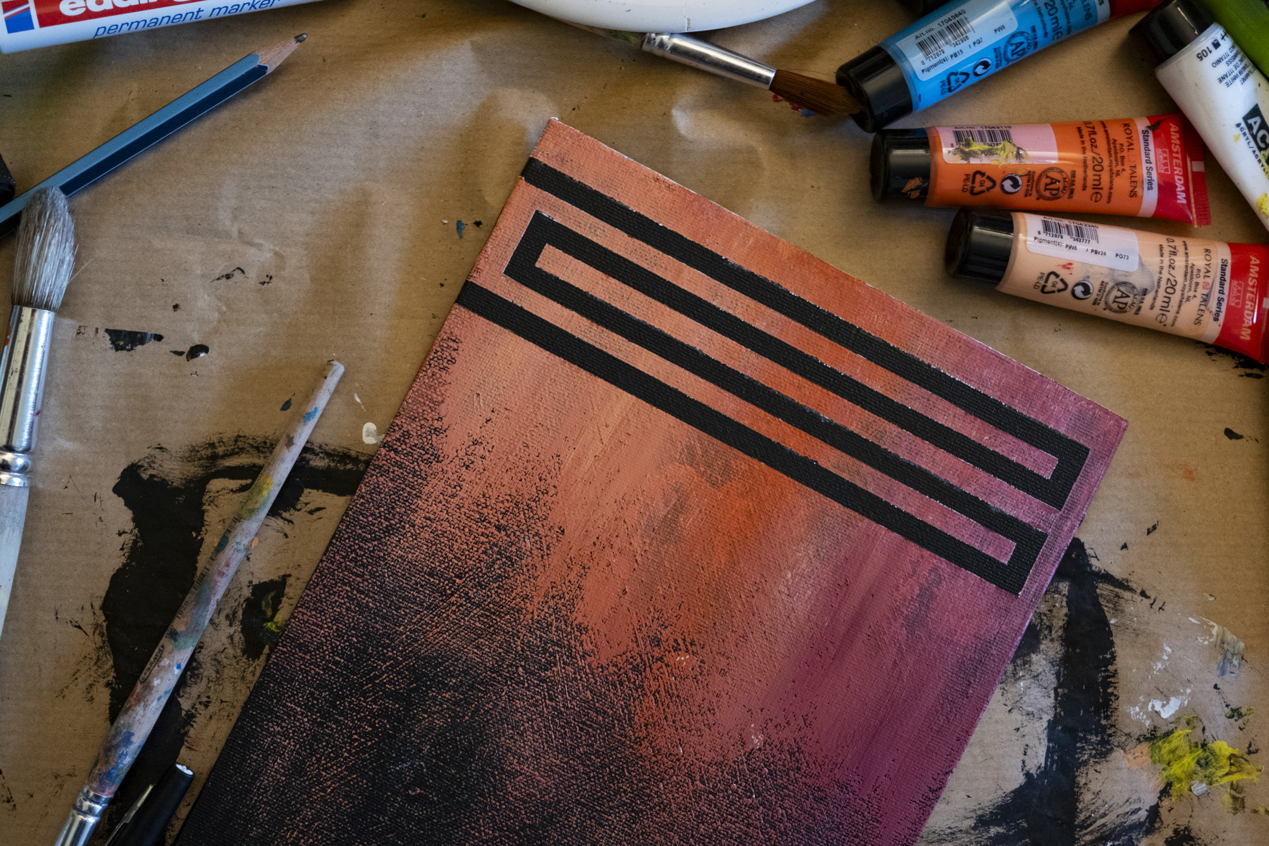 A small canvas painted in a gradient of coral orange, pink, and black, with bold black geometric lines forming a rectangular spiral near the top edge, lying on brown paper among paintbrushes, a pencil, and tubes of acrylic paint.